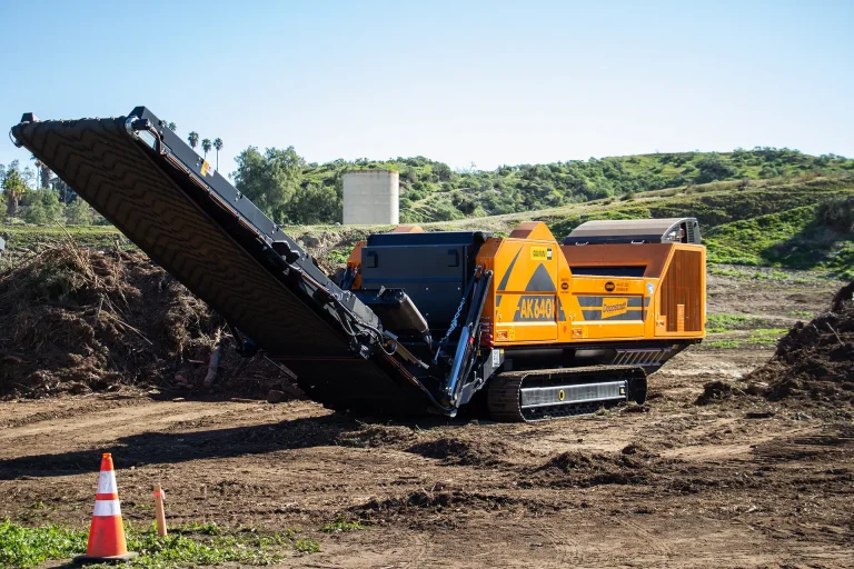 An orange Doppstant AK 640 grinder sits in front of a large pile of green waste and wood waste.