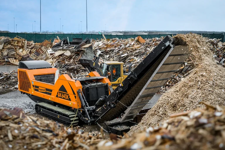 An orange Doppstant grinder sits among waste wood, grinding down pallets into wood chips.