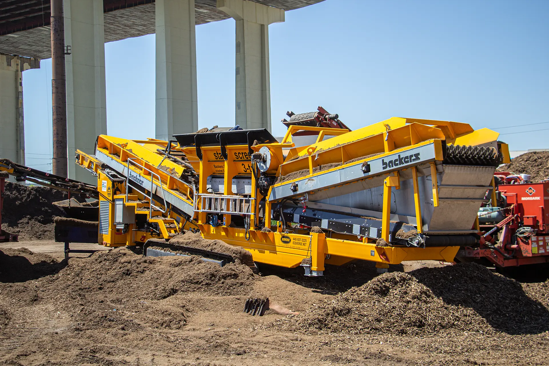 A yellow Backers star screen machine sits under a bridge as it processes and separates mulch into different sized piles.
