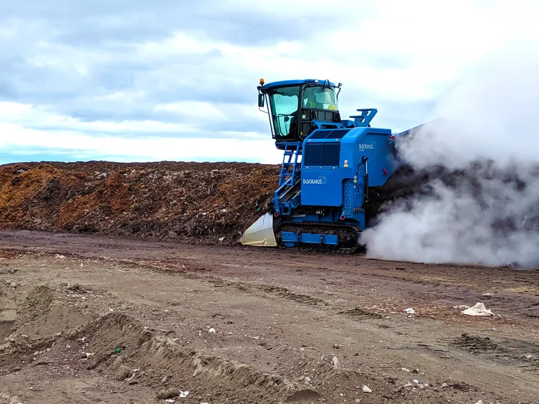 A blue BACKHUS compost turner is seen from behind as it turns a row of compost. Steam from the turned compost trails the machine.