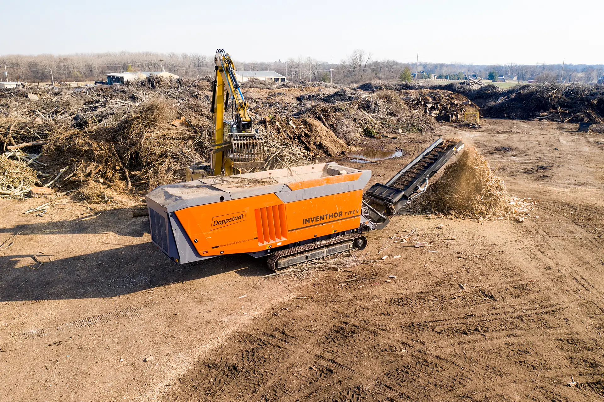 An orange Doppstant INVENTHOR 9 shredder sits in front of a large pile of green waste as a loader feeds it wood to be shredded.