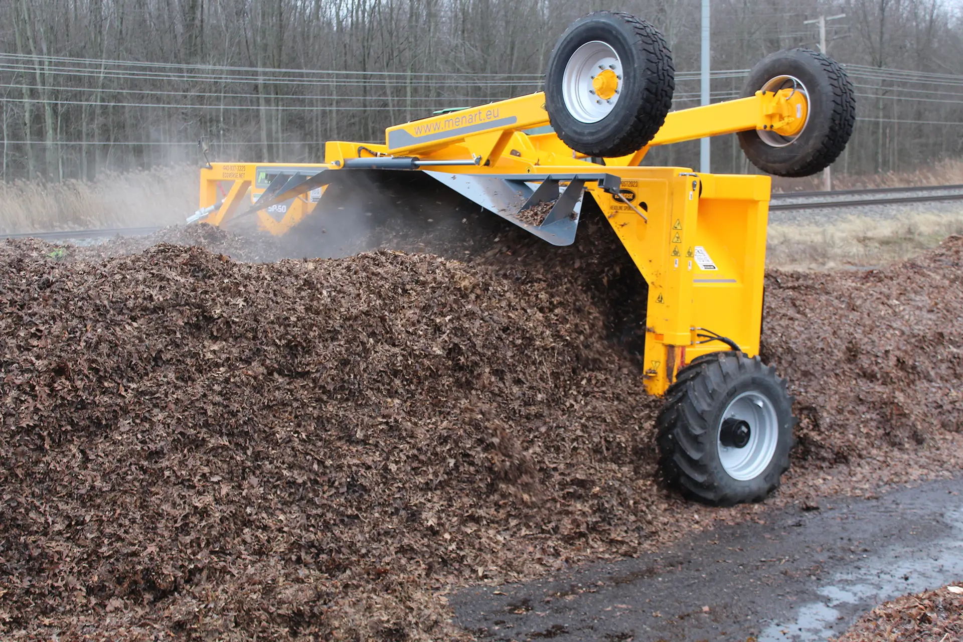 A Menart tow-behind compost turner is rolling along, turning a compost windrow.