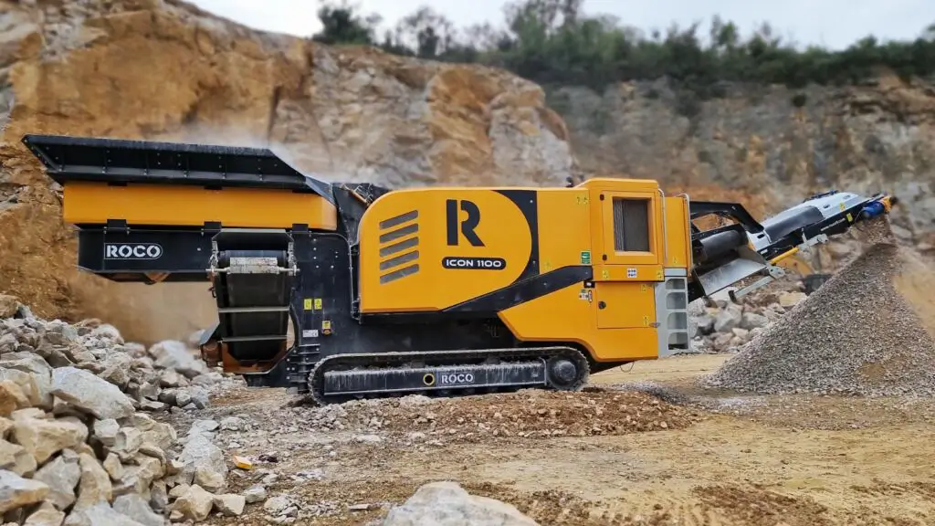 A black and amber Roco crusher is working in a quarry with crushed rocks cascading off its conveyor.