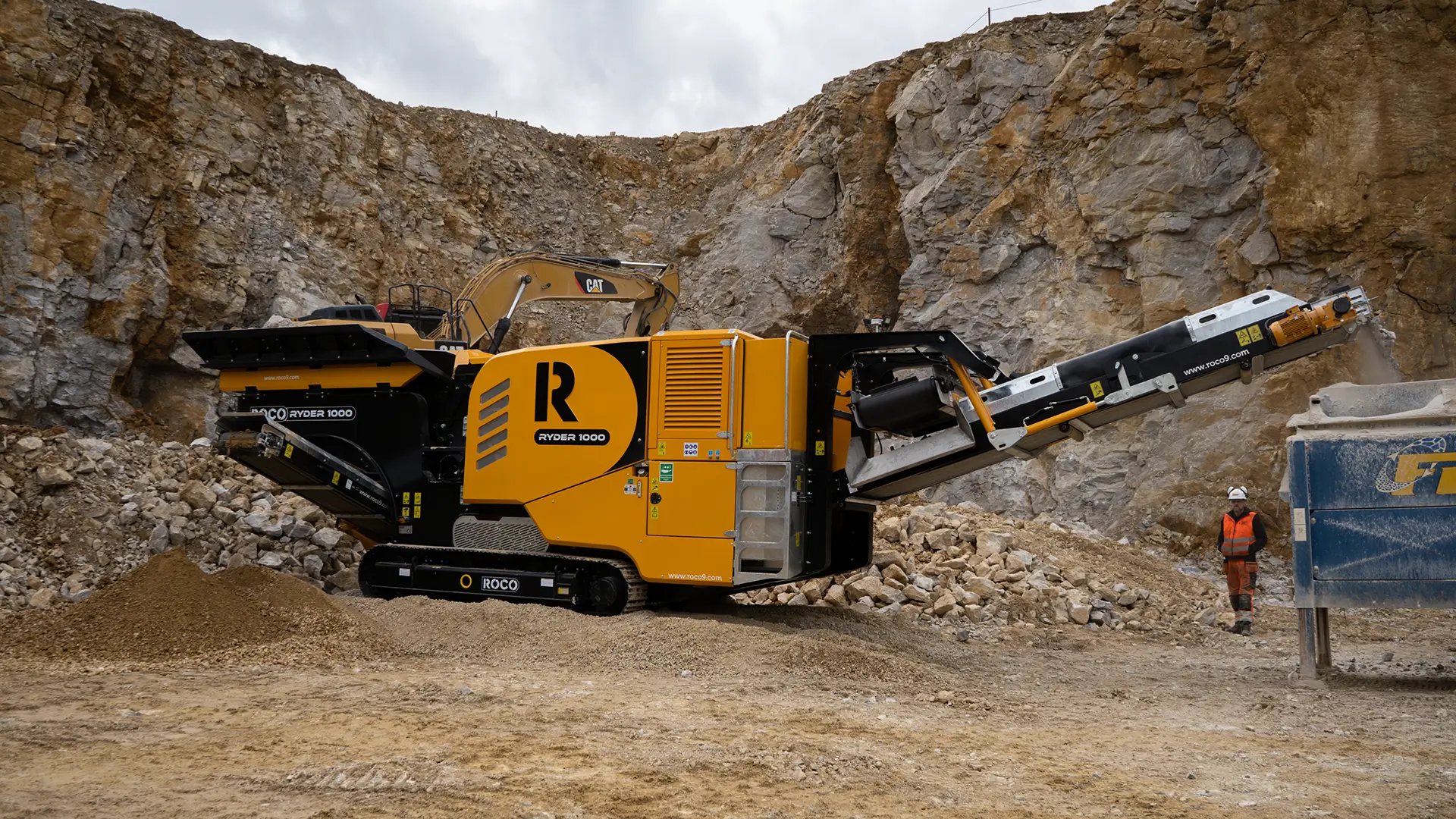 A black and amber Roco jaw crusher is shown in a high-walled quarry surrounded by crushed rocks.