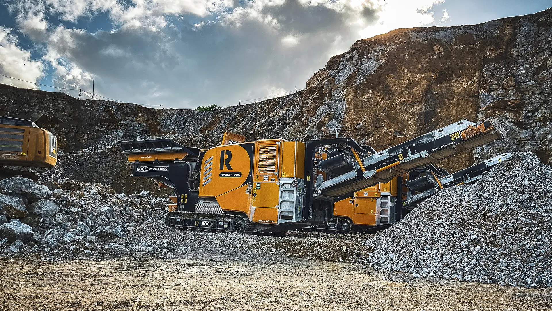 A black and amber Roco jaw crusher is working in a quarry, crushing large rocks.