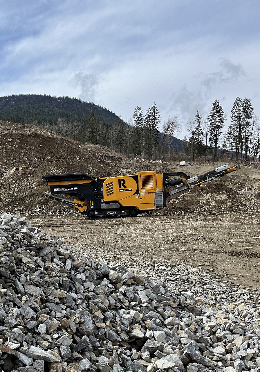 A black and amber jaw crusher sits in a quarry, surrounded by aggregate of different sizes.