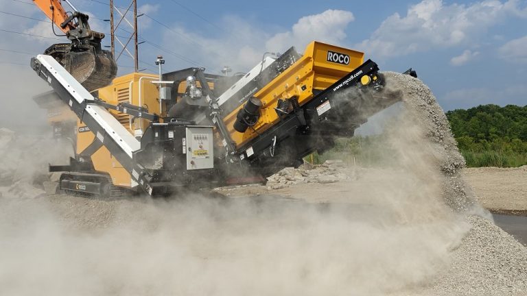 A black and amber Roco crusher is crushing recycled concrete with smaller pieces cascading off its conveyor.