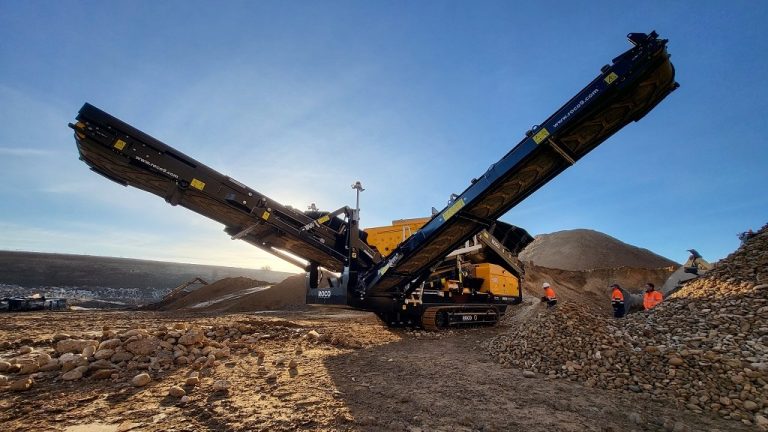 A black and amber Roco scalping screen sits among piles of rocks in the early morning, with the sun rising behind it.