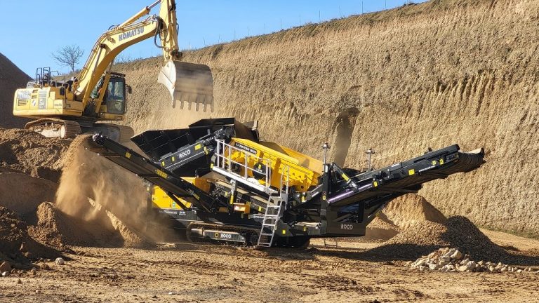 A black and amber Roco scalping screen machine sits in a high-walled quarry, separating crushed rocks into three piles of different sized material.