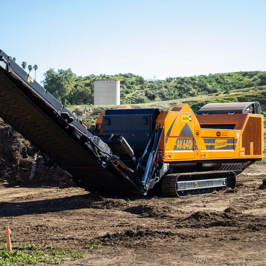An orange Doppstant AK 640 grinder sits in front of a large pile of green waste and wood waste.