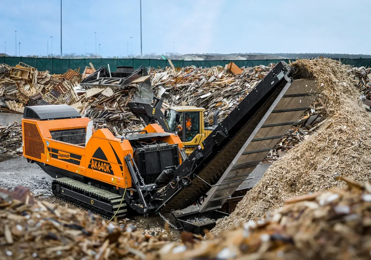 An orange Doppstant grinder sits among waste wood, grinding down pallets into wood chips.
