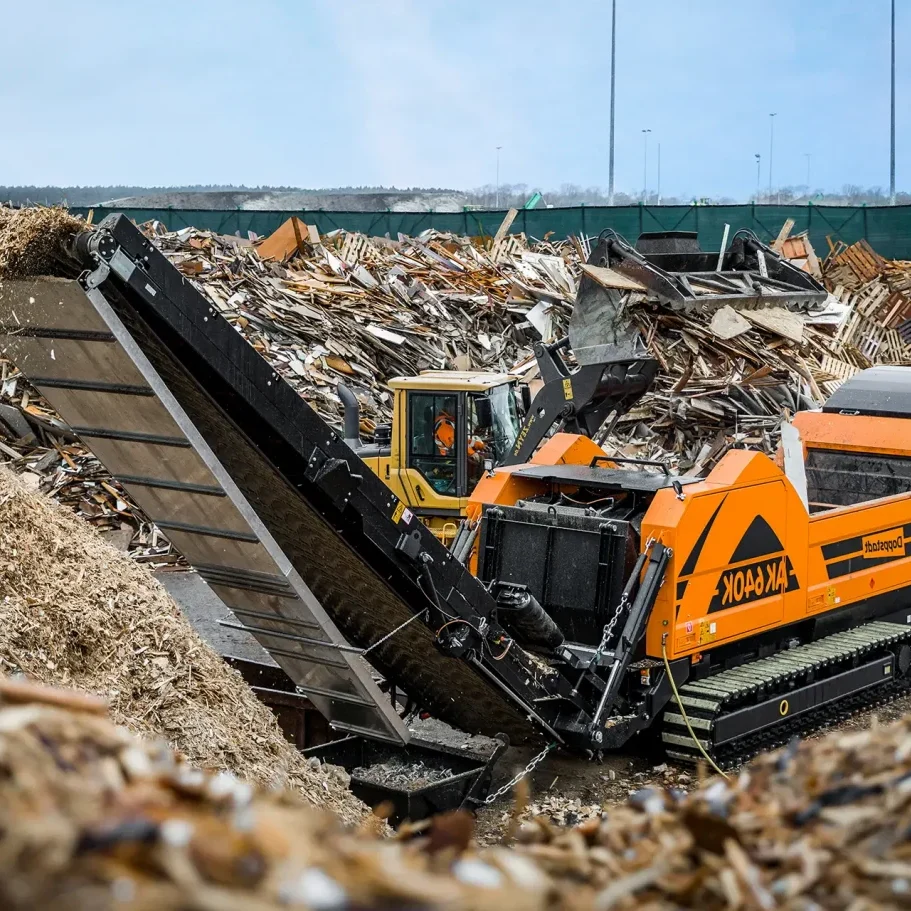 A high-speed grinder sits among piles of waste, with small ground up pieces being discharged via a conveyor.