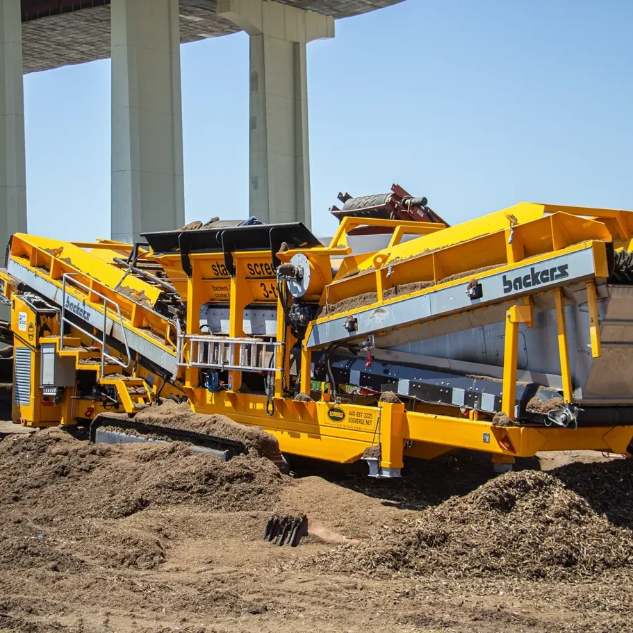 A yellow Backers star screen machine sits under a bridge as it processes and separates mulch into different sized piles.