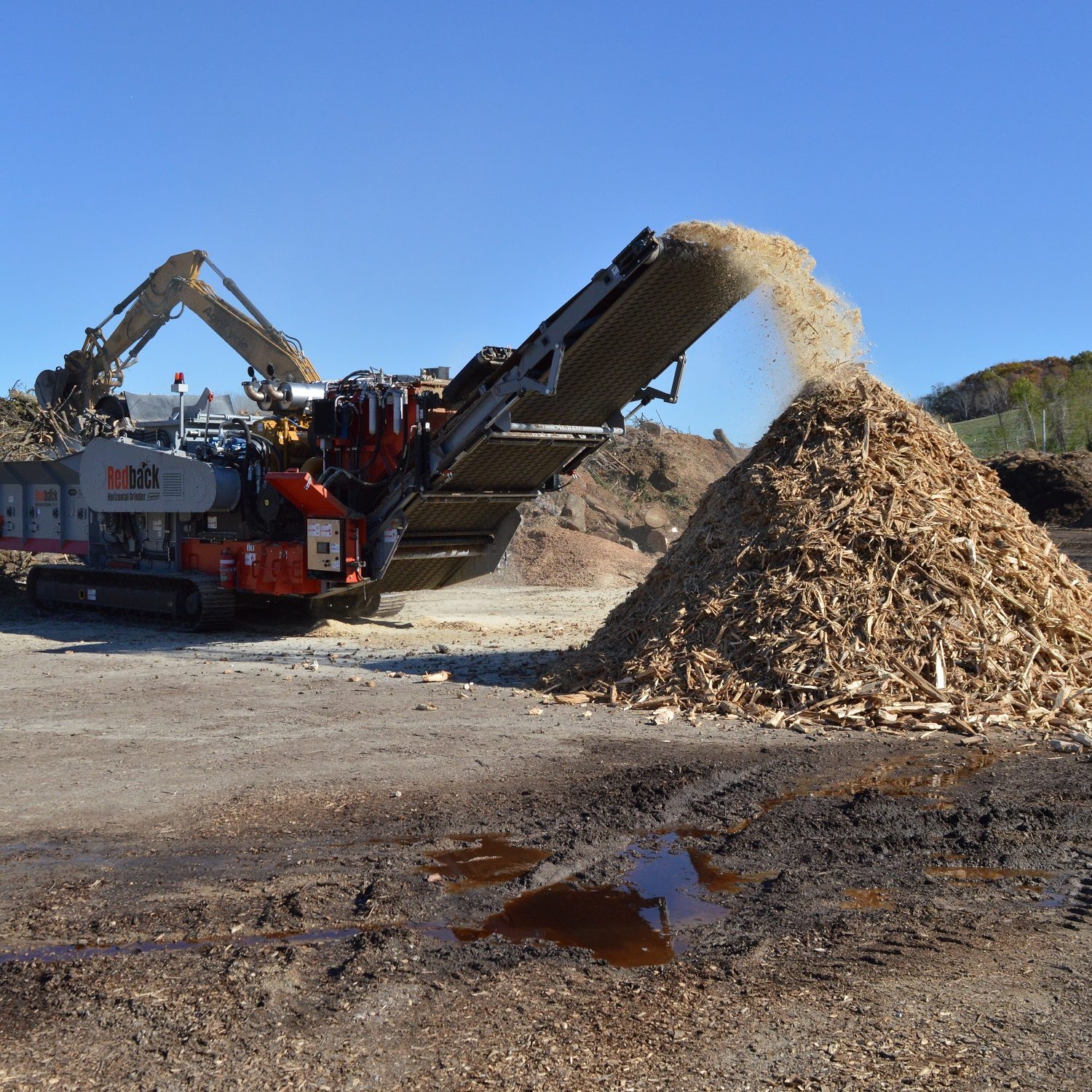 A loader adds wood to be recycled into the Redback grinder as wood chips come off its belt.