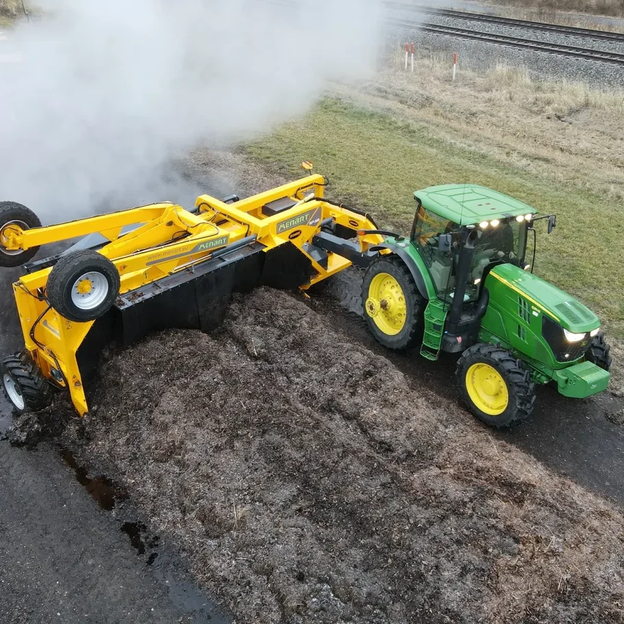 A yellow Menart tow-behind compost turner is pulled by a green tractor to turn a compost windrow.